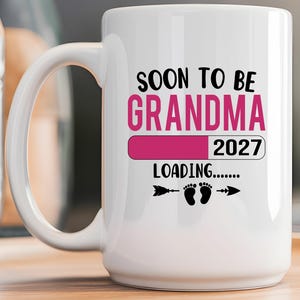 May include: A white ceramic mug featuring black and pink text that reads SOON TO BE GRANDMA, a pink loading bar, the year 2027, the word LOADING, and a graphic of two small baby footprints flanked by two black arrows pointing in opposite directions.