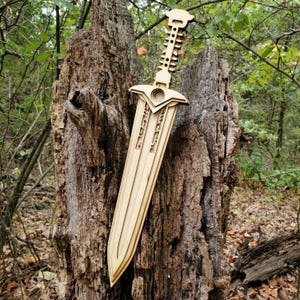 May include: A wooden sword with a detailed design, featuring a handle with a series of rectangular cutouts and a blade with engraved text. The sword is light brown and is displayed against a natural wood background.