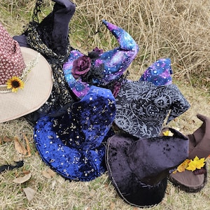 May include: Five witch hats in various styles and colors. The hats are made of velvet, with some featuring galaxy prints and others with constellations. The hats are arranged on a bed of dry grass.