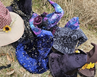 May include: Five witch hats in various styles and colors. The hats are made of velvet, with some featuring galaxy prints and others with constellations. The hats are arranged on a bed of dry grass.