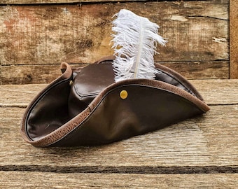 May include: A brown leather pirate hat with a white feather sticking out of the top. The hat is sitting on a wooden surface.