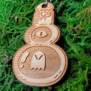 May include: A wooden keychain with a whimsical design. The keychain features a layered design with a ghost, an eye, and other decorative elements. The keychain is made of light-colored wood and has a metal ring for attaching keys.