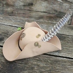 May include: A tan felt tricorn hat with a black band and decorative metal gears. A single white and gray feather is attached to the hat.