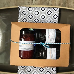 May include: A gift box with a window showing three jars of jam. The jars are labeled "Strawberry Rhubarb Jam", "Sour Cherry Jam", and "Juniper Berry Jam". The box is tied with blue and white twine.