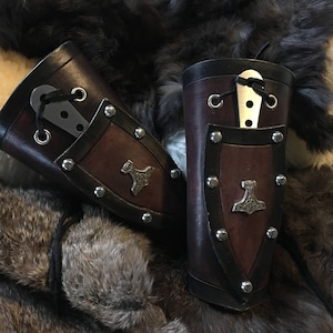 May include: Two brown leather arm bracers with metal accents. Each bracer has a small metal hammer design and a slot for a knife. The bracers are on a brown and white fur background.