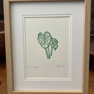 Op de afbeelding: Ingelijste print met een groene illustratie van tarobladen op een witte achtergrond. Het kunstwerk is gesigneerd en bevat de tekst "Taro patch". De print is geplaatst in een witte passe-partout en een lichtbruine houten lijst.