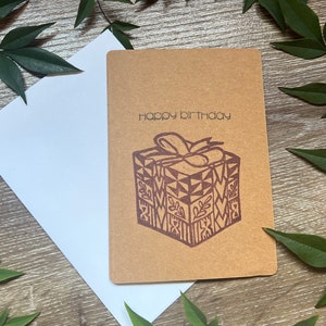 Pode incluir: Um cartão de aniversário castanho com as palavras "happy birthday" impressas no topo. Uma caixa de presente com um laço é impressa abaixo do texto. Um envelope branco está atrás do cartão. Folhas verdes no fundo.