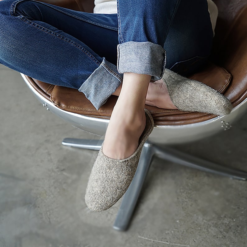 May include: A pair of gray wool slippers with a closed toe and a slip-on design. The slippers are worn by a person sitting in a chair.