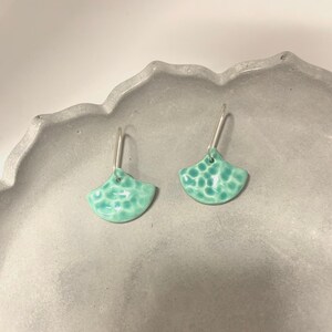 Può includere: Un paio di orecchini in ceramica turchese a forma di ventaglio con superficie strutturata. Gli orecchini hanno ganci color argento. Gli orecchini sono esposti su una superficie grigia strutturata.