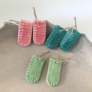 Op de afbeelding: Vier paar keramische oorbellen in roze, teal en groen. Elke oorbel heeft een rechthoekige vorm met een textuurpatroon en zilverkleurige oorhaken. De oorbellen worden gepresenteerd op een grijze, getextureerde ondergrond.