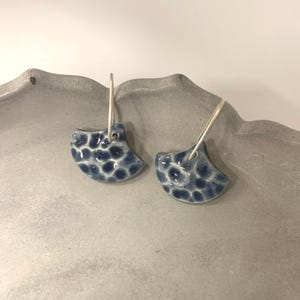 Può includere: Un paio di orecchini in ceramica a forma di ventaglio. Gli orecchini sono blu scuro con un motivo strutturato e maculato, appesi a cerchi argentati. Sono esposti su una superficie grigia.