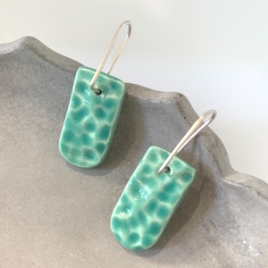 May include: Two turquoise ceramic earrings with a textured pattern. The rectangular earrings have rounded edges and are suspended from silver-coloured ear wires. The earrings are presented on a grey, textured surface.