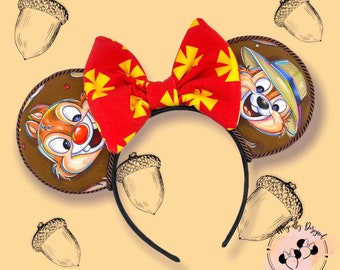Puede incluir: Diadema con dos orejas circulares de tela marrón con ilustraciones de Chip y Chop. Un gran lazo de tela roja con un patrón de estrellas amarillas se sitúa entre las orejas. La diadema es negra y fina, decorada con ilustraciones de bellotas.