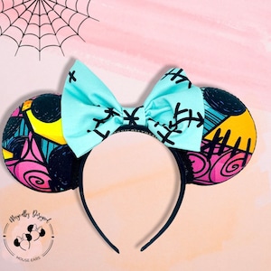 Puede incluir: Diadema de orejas de Minnie Mouse con un lazo azul claro con detalles de costuras negras. Las orejas tienen un diseño de patchwork colorido con formas negras, amarillas, rosas y azules. La diadema es negra y el fondo es rosa.