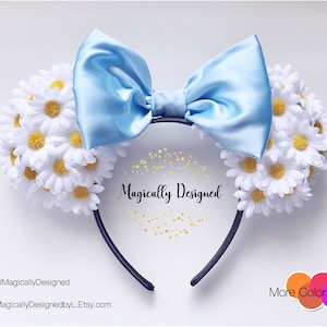 May include: Light blue satin bow headband with white daisy flower accents. The headband is black and says "Magically Designed".