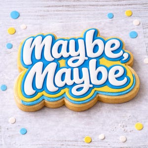 Pu&ograve; includere: Un biscotto decorato con le parole "Maybe, Maybe" in lettere bianche su sfondo blu e giallo. Il biscotto &egrave; circondato da zuccherini blu, gialli e bianchi su una superficie bianca.