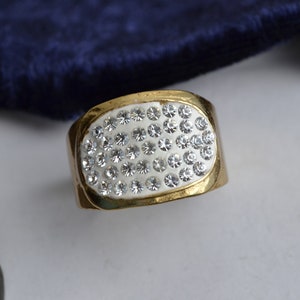 May include: A gold ring with a white oval-shaped setting encrusted with small clear rhinestones.