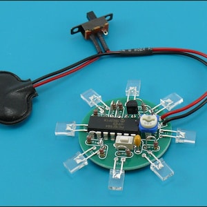 May include: A small, round green circuit board with eight clear LED lights arranged in a circle. It has a black battery holder, a switch, and various electronic components. Red and black wires connect the components.