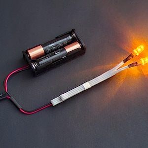 Peut inclure: Un support de batterie noir avec deux piles AA, relié par des fils rouges et noirs à un interrupteur et à deux lumières LED jaunes brillantes. Les lumières émettent une lueur chaude et orange sur un fond sombre.
