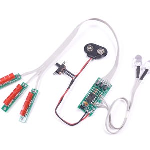 May include: Electronic components on a white background. The image shows a battery, circuit boards, red and green components, and LED lights. Wires connect the parts, suggesting a DIY electronics project or kit.