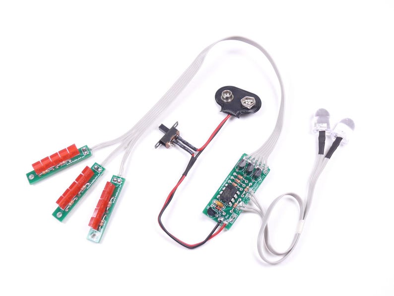 May include: Electronic components on a white background. The image shows a battery, circuit boards, red and green components, and LED lights. Wires connect the parts, suggesting a DIY electronics project or kit.