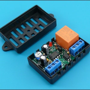 May include: A black plastic enclosure with a circuit board inside. The board has a red LED, a small orange relay, and several blue terminal blocks. The enclosure has a removable lid with a grid pattern.