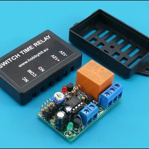 Op de afbeelding: Een Switch Time Relay printplaat met een zwarte plastic behuizing en een apart zwart deksel. De printplaat heeft diverse elektronische componenten, waaronder een oranje relais, blauwe klemmenblokken en een groene LED. De tekst "SWITCH TIME RELAY" is zichtbaar.