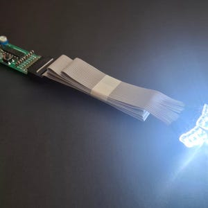 May include: A green electronic circuit board with a red wire, connected to a grey ribbon cable that leads to three illuminated white LED lights. The circuit board features a small blue component and a black integrated chip on its surface.