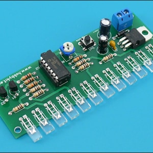 Puede incluir: Una placa de circuito verde con varios componentes electrónicos, incluyendo resistencias, condensadores y circuitos integrados. La placa presenta componentes de plástico transparente y el texto "www.hobbykit.eu" sobre un fondo azul.