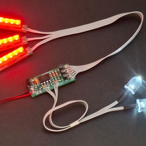 May include: Electronic components featuring three red LED light strips, a circuit board, and two white LED lights. The components are connected by white ribbon cables and black and red wires, set against a dark background.