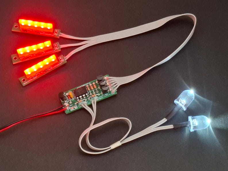 May include: Electronic components featuring three red LED light strips, a circuit board, and two white LED lights. The components are connected by white ribbon cables and black and red wires, set against a dark background.