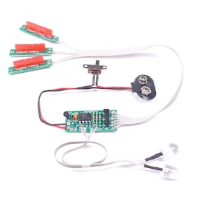 May include: Electronic circuit components on a white background. The components include three small circuit boards with red connectors, a switch, a 9-volt battery connector, a small circuit board, and two white LED lights.