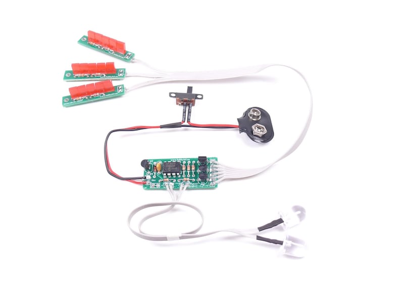 May include: Electronic circuit components on a white background. The components include three small circuit boards with red connectors, a switch, a 9-volt battery connector, a small circuit board, and two white LED lights.