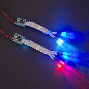 Könnte beinhalten: Zwei elektronische Leiterplatten mit angeschlossenen roten Drähten und Flachbandkabeln, die zu mehreren kleinen LED-Leuchten führen. Die LEDs strahlen helles blaues und rotes Licht aus und erzeugen einen fokussierten Beleuchtungseffekt.