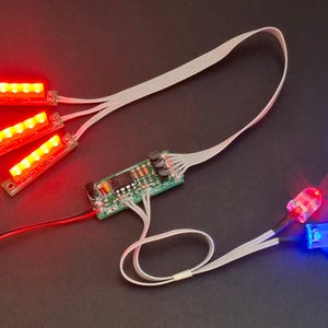 May include: Electronic components featuring red and blue LEDs, along with three red LED light strips. The components are connected to a green circuit board with various electronic elements. White ribbon cables and red and black wires are also visible.