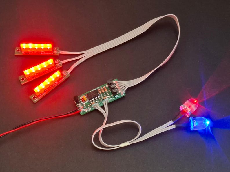 May include: Electronic components featuring red and blue LEDs, along with three red LED light strips. The components are connected to a green circuit board with various electronic elements. White ribbon cables and red and black wires are also visible.