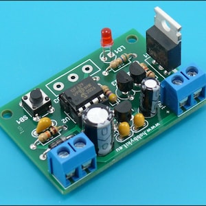 May include: A green circuit board with electronic components, including a red LED, a button, a transistor, and a capacitor. The board has a blue terminal block and the text "SB7" and "100" printed on it.