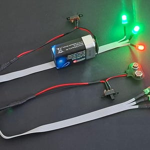 May include: Two electronic circuit kits, each featuring a 9-volt alkaline battery connected to a black slide switch and three LED lights. One kit has two green and one red illuminated LEDs, while the other has two green and one red unlit LEDs.