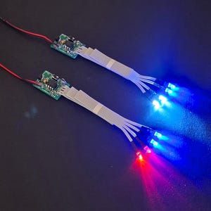 Könnte beinhalten: Zwei elektronische Leiterplatten mit Flachbandkabeln, die zu mehreren Leuchtdioden führen. Die LEDs leuchten in hellem Blau und Rot und sind über dünne rote und schwarze Drähte mit kleinen Netzschaltern an den Enden verbunden.
