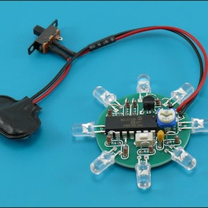 May include: A green circuit board featuring eight clear LED lights in a circular arrangement. It includes a black battery holder, a switch, and various electronic components. Red and black wires connect the components.