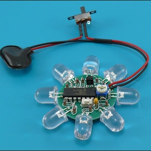 May include: A green circuit board with eight clear LED lights arranged in a circular pattern. The board has various electronic components, including a chip and a potentiometer. Black and red wires connect to a black battery holder and a small switch.