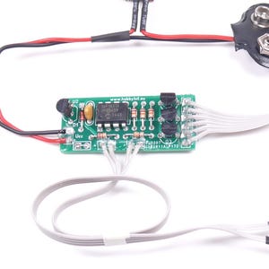 May include: A small green circuit board with various electronic components, including resistors and an integrated circuit. It is connected to a 9-volt battery connector with black and red wires, and white wires with black heat shrink tubing. The image is on a white background.
