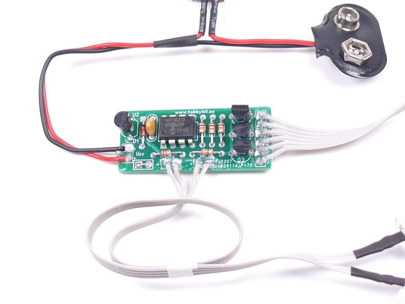 May include: A small green circuit board with various electronic components, including resistors and an integrated circuit. It is connected to a 9-volt battery connector with black and red wires, and white wires with black heat shrink tubing. The image is on a white background.