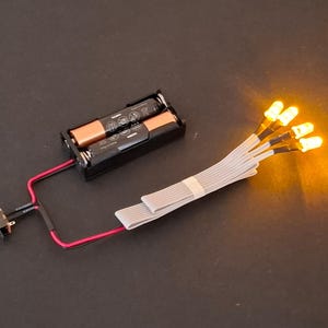 Peut inclure: Un support de batterie noir avec deux piles, relié par des fils rouges à un interrupteur et un faisceau de fils gris avec cinq lumières LED jaunes. Les lumières sont allumées, émettant une lueur chaude.