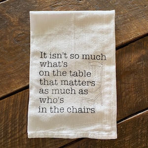 May include: White tea towel with black text that reads "It isn't so much what's on the table that matters as much as who's in the chairs". A simple line drawing of a chair is included in the text.