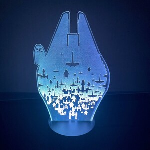 May include: A blue and white acrylic light sculpture of the Millennium Falcon spaceship from Star Wars. The ship is surrounded by smaller spaceships, creating a detailed and intricate design.