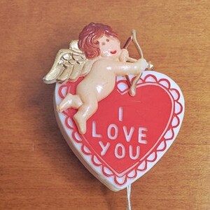 May include: A red heart-shaped ornament with a white border and the words "I LOVE YOU" in white lettering. A small cherub with gold wings and a bow and arrow is attached to the top of the heart. The ornament is hanging from a white string with a red bell at the end.