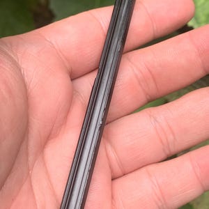 May include: A black tourmaline crystal wand, held in a hand. The wand is a dark, glossy black with vertical ridges along its length. The crystal is a natural, raw form, showcasing its unique texture and shape.