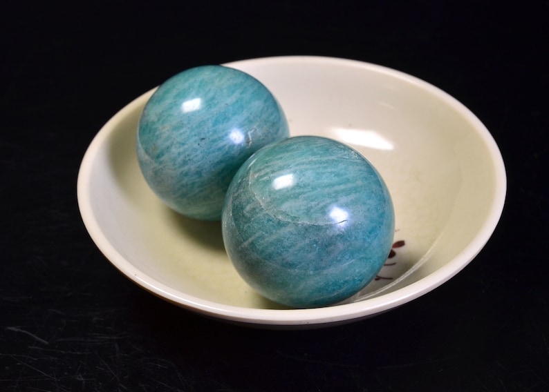 May include: Two smooth, green, round, polished, gemstone spheres in a white bowl. The spheres are made of amazonite.