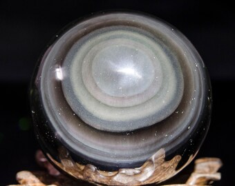May include: A polished obsidian sphere with concentric rings of gray, white, and black. The sphere is sitting on a wooden stand.